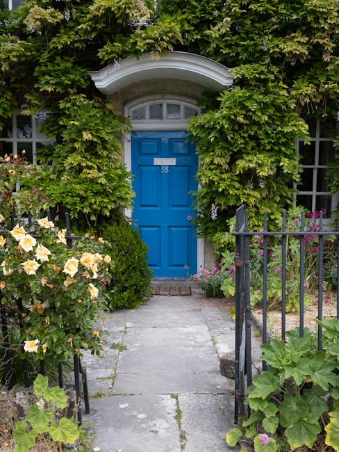 A charming front garden with a narrow, paved stone pathway leading to a vibrant blue door set within a white arched doorway, partially obscured by lush green foliage and climbing plants. The garden fe