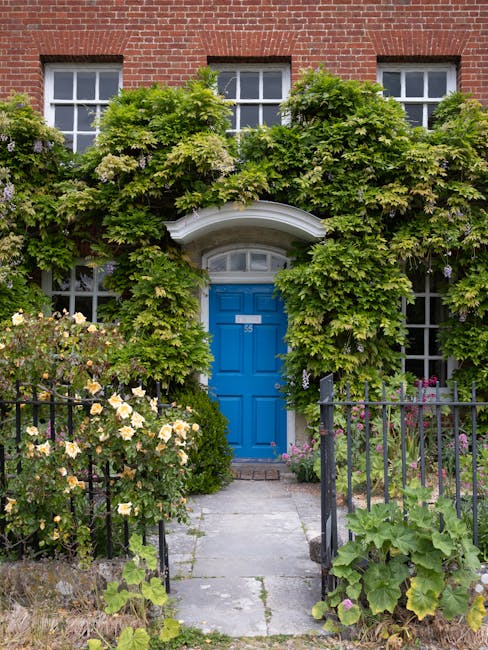 A charming front garden with a narrow, paved stone pathway leading to a vibrant blue door set within a white arched doorway, partially obscured by lush green foliage and climbing plants. The garden fe