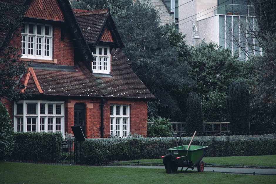 A quaint red-brick house with a steep, tiled roof and white-framed windows, surrounded by a well-maintained garden with neatly trimmed hedges and lush greenery. In the foreground, a green garden wheel